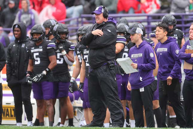 A white man with a purple hat and black headphones, wearing all black, stands with his arms folded on the sidelines of a football game, flanked by players and staff dressed in black and purple.