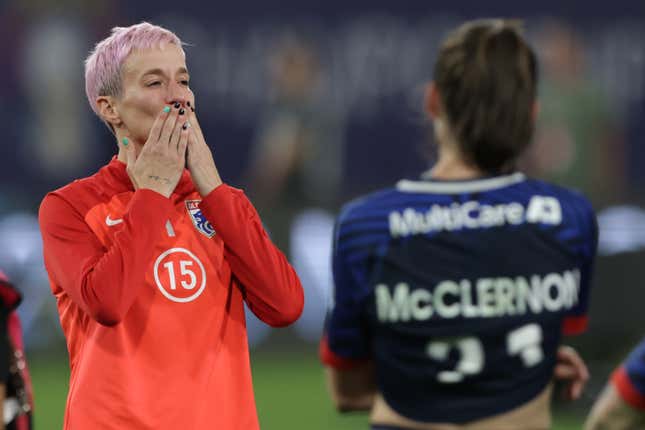 A white woman with short pink hair and a red Nike warmup blows kisses to the crowd while standing on a soccer pitch.